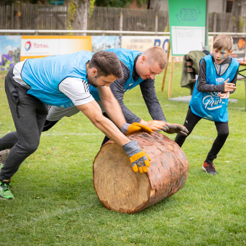 Zwei Teilnehmer des Mitarbeiterfests in blauen Trikots rollen einen kurzen Baumstumpf vor sich her. Das Kind eines Mitarbeiters feuert sie an.