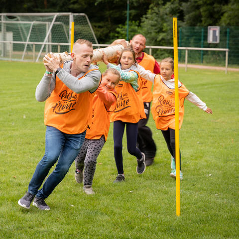 Sportfest-Teilnehmer, zwei Männer und drei Kinder in orangen Team-Trikots, tragen gemeinsam einen Baumstamm und laufen mit diesem um Hindernisse herum.