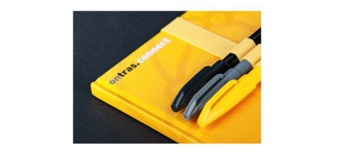 Ein gelbes Notizbuch mit dem Aufdruck ONTRAS connect. Am Notizbuch sind drei Stifte mit einem dekorativen Stifthalter befestigt.