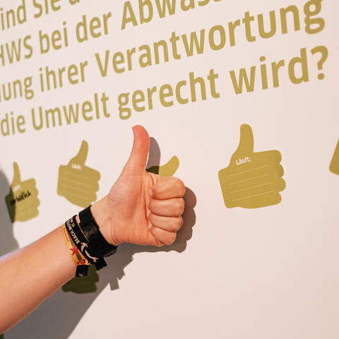 Eine Hand mit Daumen hoch wird vor einer Pinnwand mit grünen Daumen-Hoch-Stickern präsentiert.