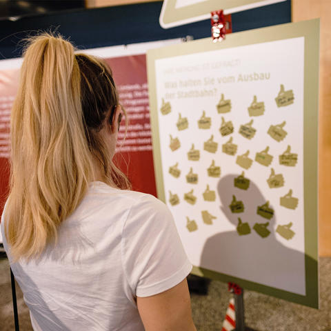Ein junge Frau mit blonden Haaren schaut auf eine Pinnwand mit grünen Daumen-Hoch-Stickern.