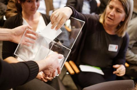 Mitarbeiter werfen ihre Fragen an die Geschäftsführer auf Papier geschrieben in einen transparenten Plexiglaswürfel.