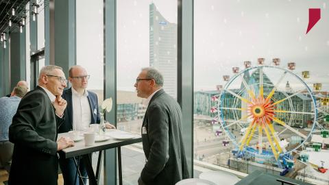 Drei Geschäftsführer der SWH Gruppe diskutieren angeregt miteinander. Die Location der Führungskräftetagung bietet Ausblick auf den Augustusplatz mit MDR Tower, den Uni-Campus und das Paulinum sowie das Gewandhaus zu Leipzig.