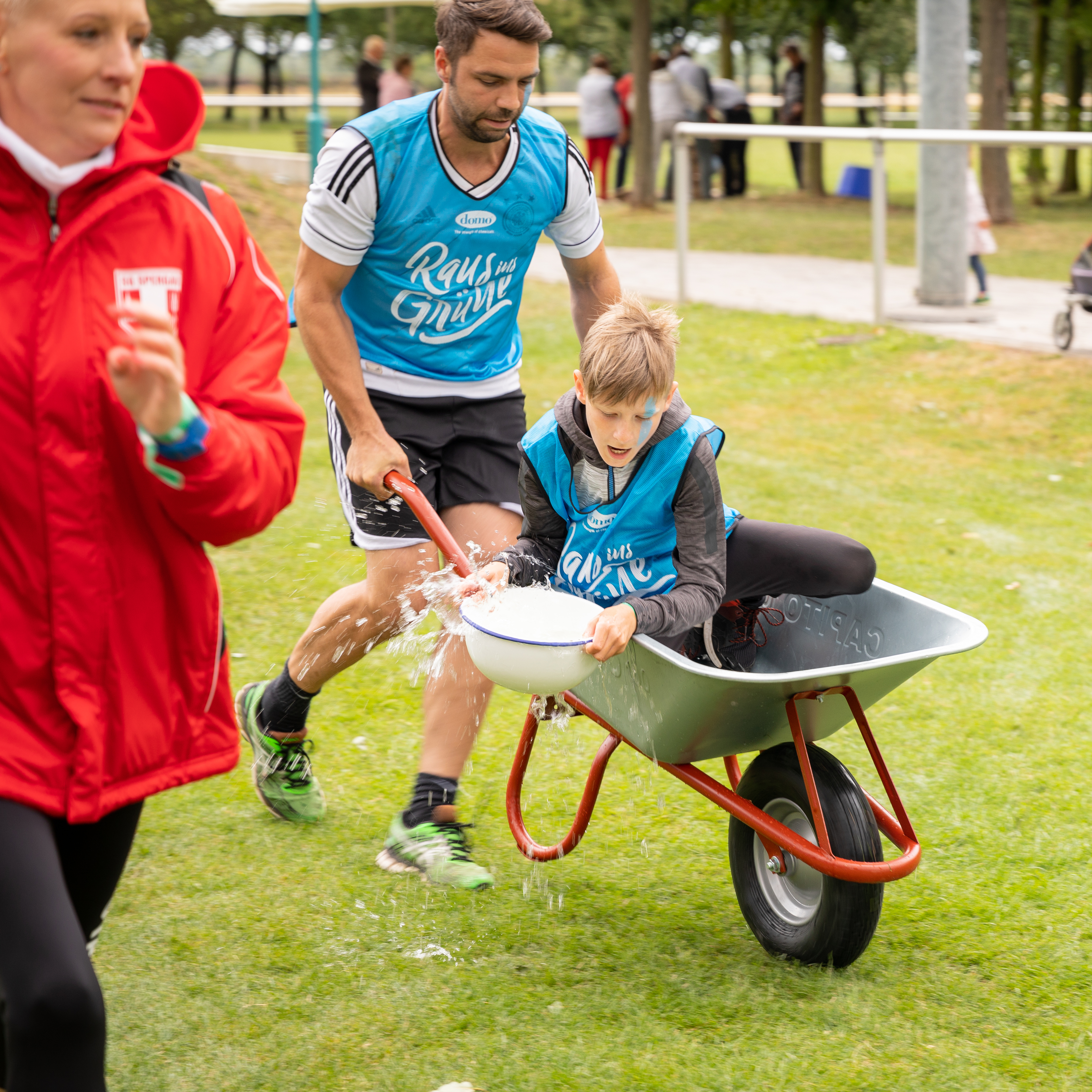 Ein Kind sitzt in einer Schubkarre und versucht dabei, eine Wasserschüssel gerade zu halten. Die Schubkarre wird von einem Erwachsenen durch einen Parkour geschoben.