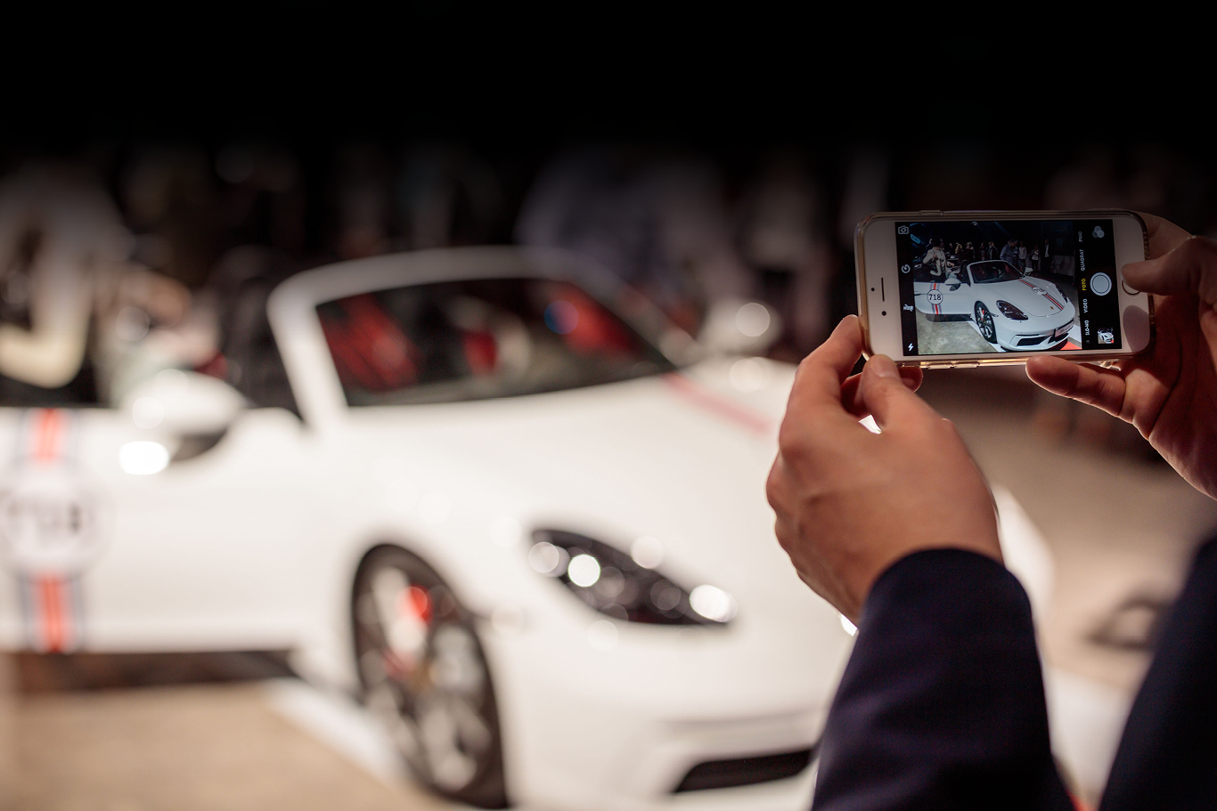 Im Hintergrund ist verschwommen ein Boxster 718 von Porsche zu sehen, welcher im Outdoorbereich der Eventlocation mit einem Handy fotografiert wird.