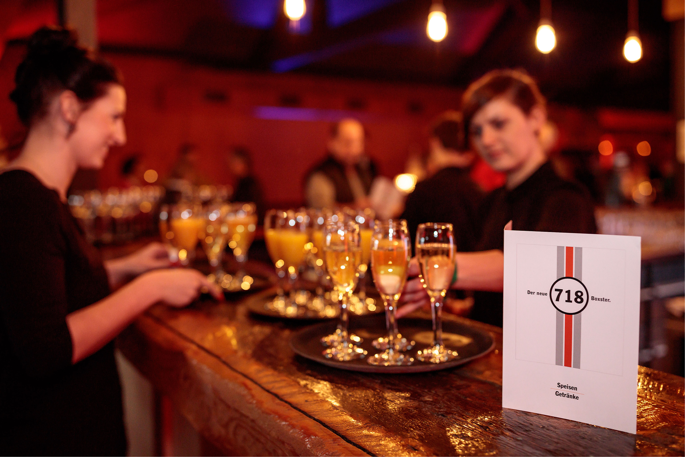 Zwei lächelnde Servicekräfte vom Event-Catering schenken Sekt und Orangensaft aus. Im Vordergrund steht eine Menükarte mit dem Boxster 718-Signet.