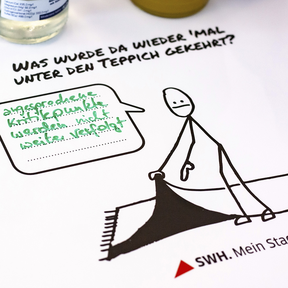 In die Sprechblase der Karikatur, welche Probleme unter den Teppich gekehrt würden, schreibt ein Mitarbeiter „angesprochene Kritikpunkte“.