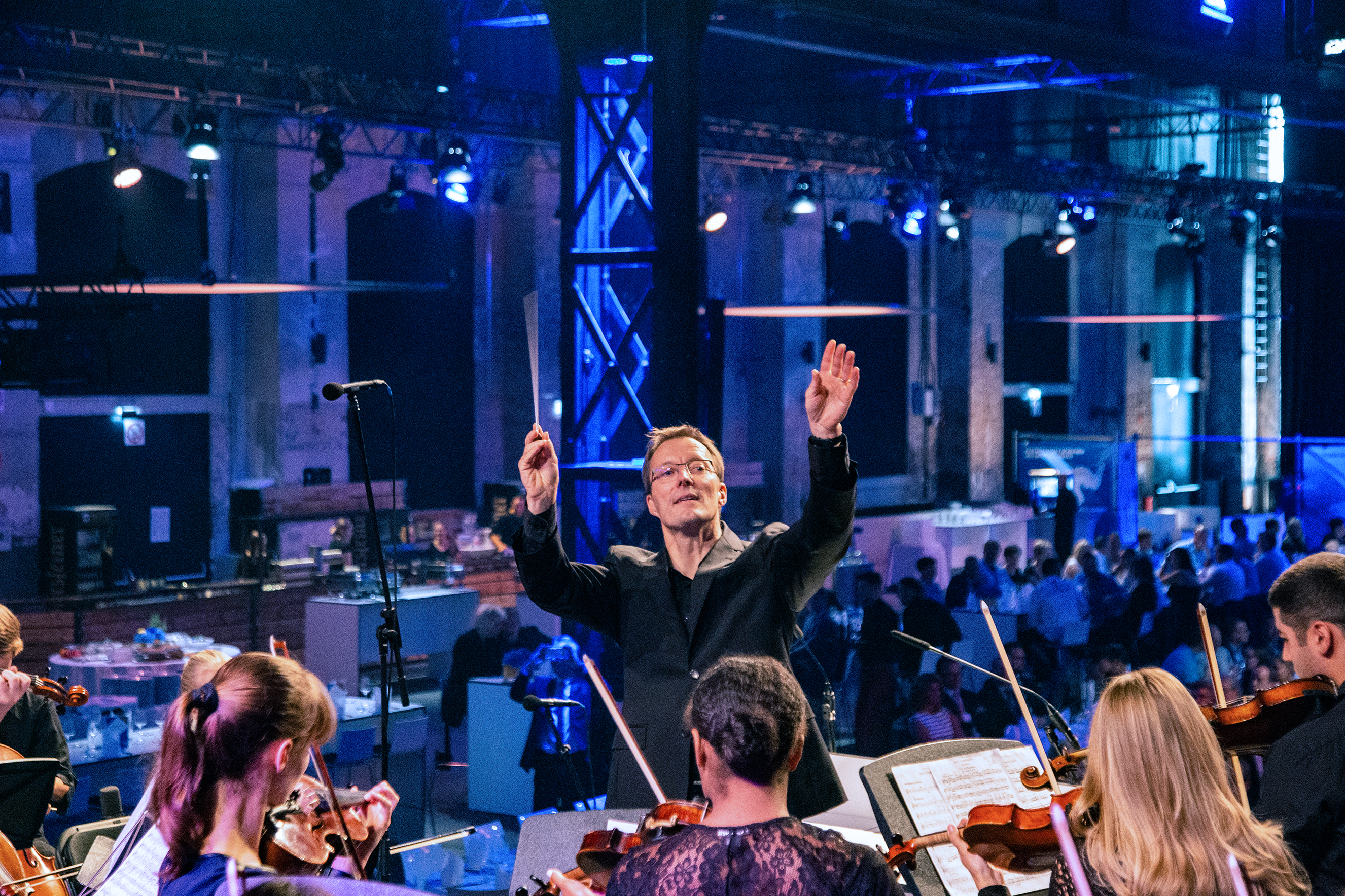 Im Vordergrund steht der Dirigent des Jugendsinfonieorchesters. Dahinter sitzen die Gäste der Jubiläumsfeier und genießen die musikalische Untermalung.