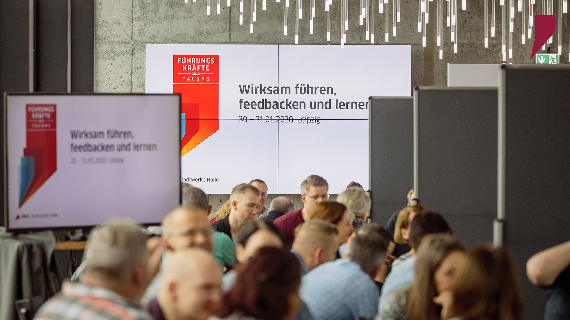 Blick in den Tagungsraum im Felix Leipzig. Die Führungskräfte sitzen in ihren Workshopgruppen zusammen. Im Hintergrund sieht man das Key Visual der Veranstaltung und das Motto: „Wirksam führen, feedbacken und lernen“.